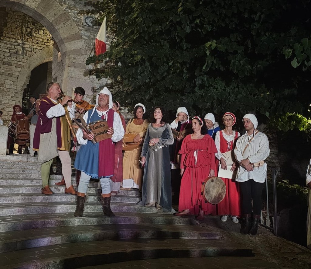 Rappresentazione storica durante l'Agosto Corcianese, con partecipanti in costumi medievali sul gradino di una scala, mentre suonano strumenti e cantano.