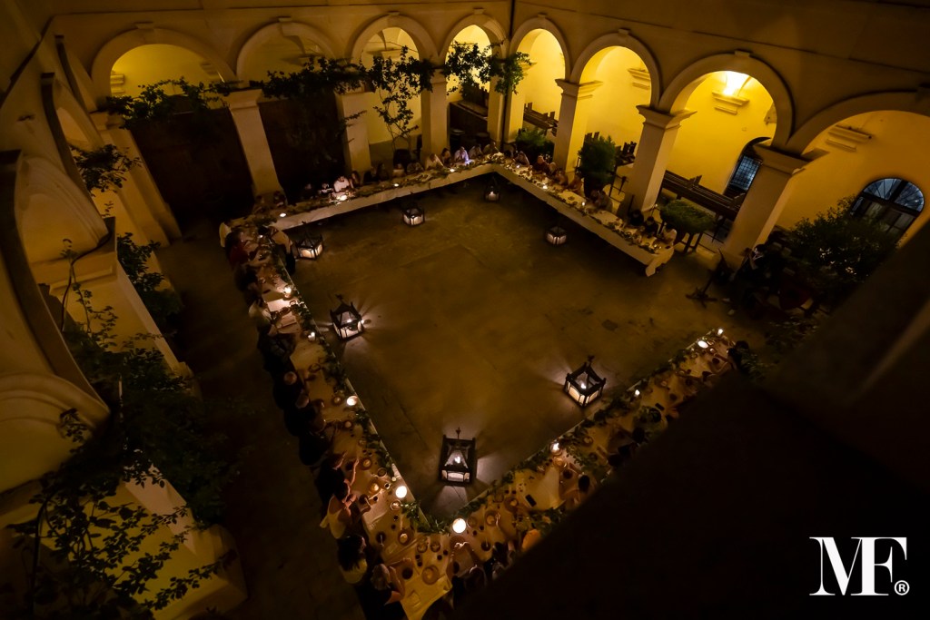 Veduta aerea di un banchetto medievale allestito in un cortile. Tavoli a forma di U disposti intorno a una pavimentazione centrale, illuminati da lanterne e decorati con piante. Diverse persone sedute attorno ai tavoli, immersi in un'atmosfera conviviali.