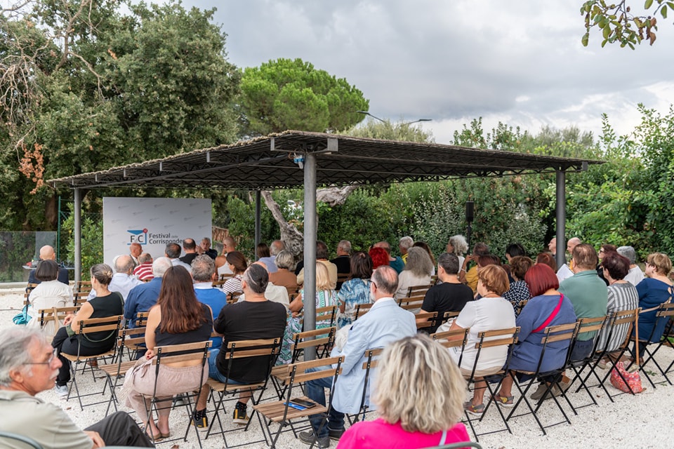 Audience seated at the Festival delle Corrispondenze, gathered under a covered area surrounded by greenery.