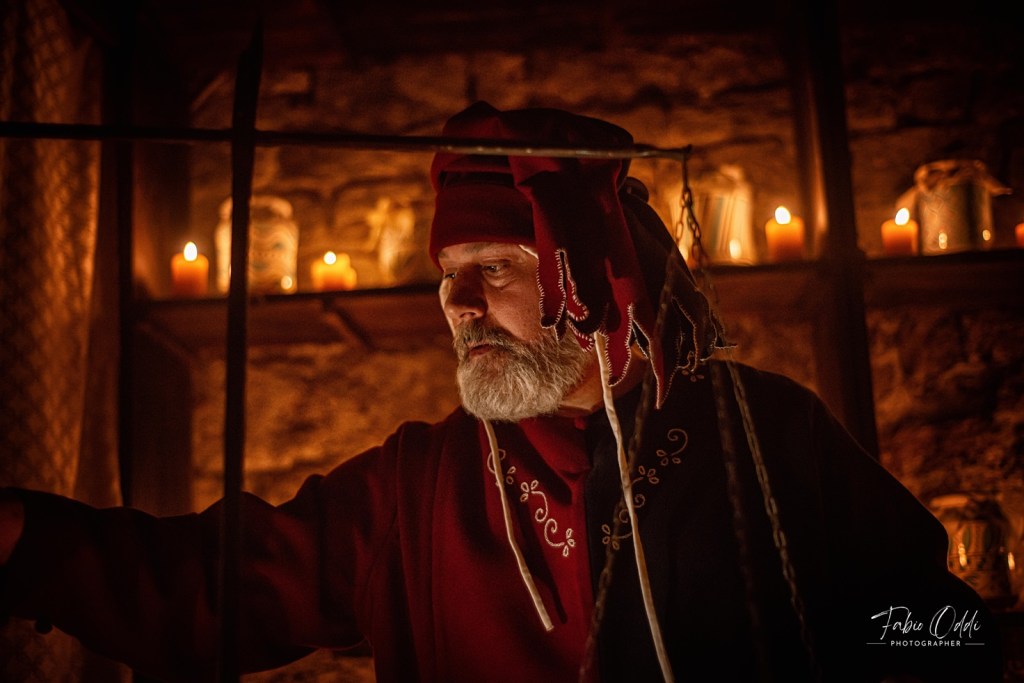 Un uomo anziano vestito in abiti medievali rossi, con una barba grigia, mentre interagisce con oggetti in un ambiente illuminato da candele.
