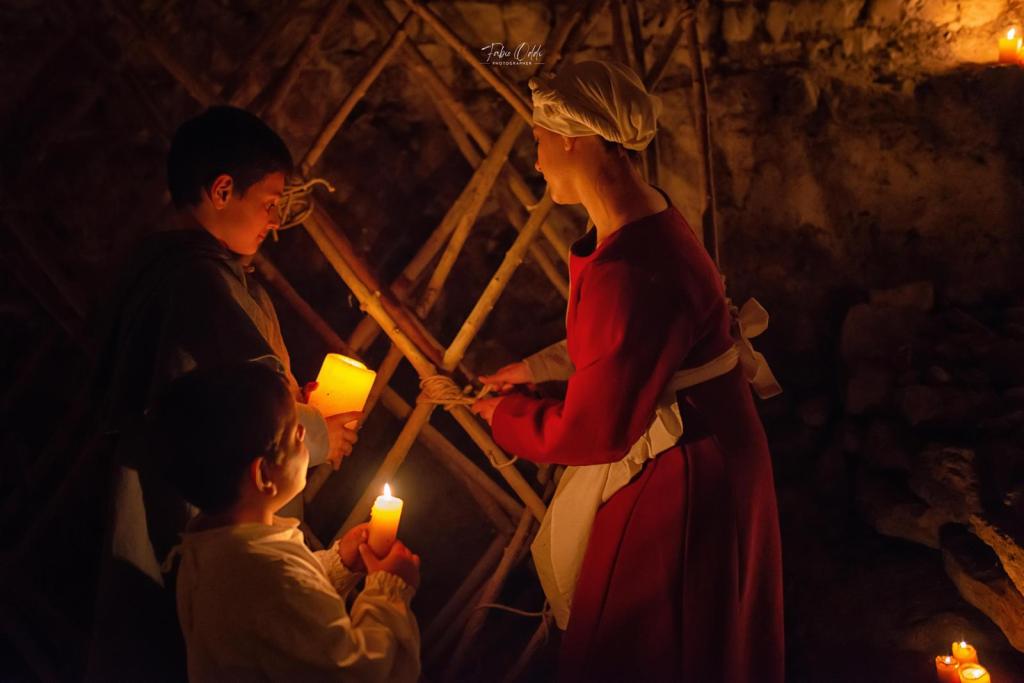 Due bambini e una donna vestita in abiti medievali, all'interno di un ambiente illuminato da candele, mentre si scambiano candele e conversano.