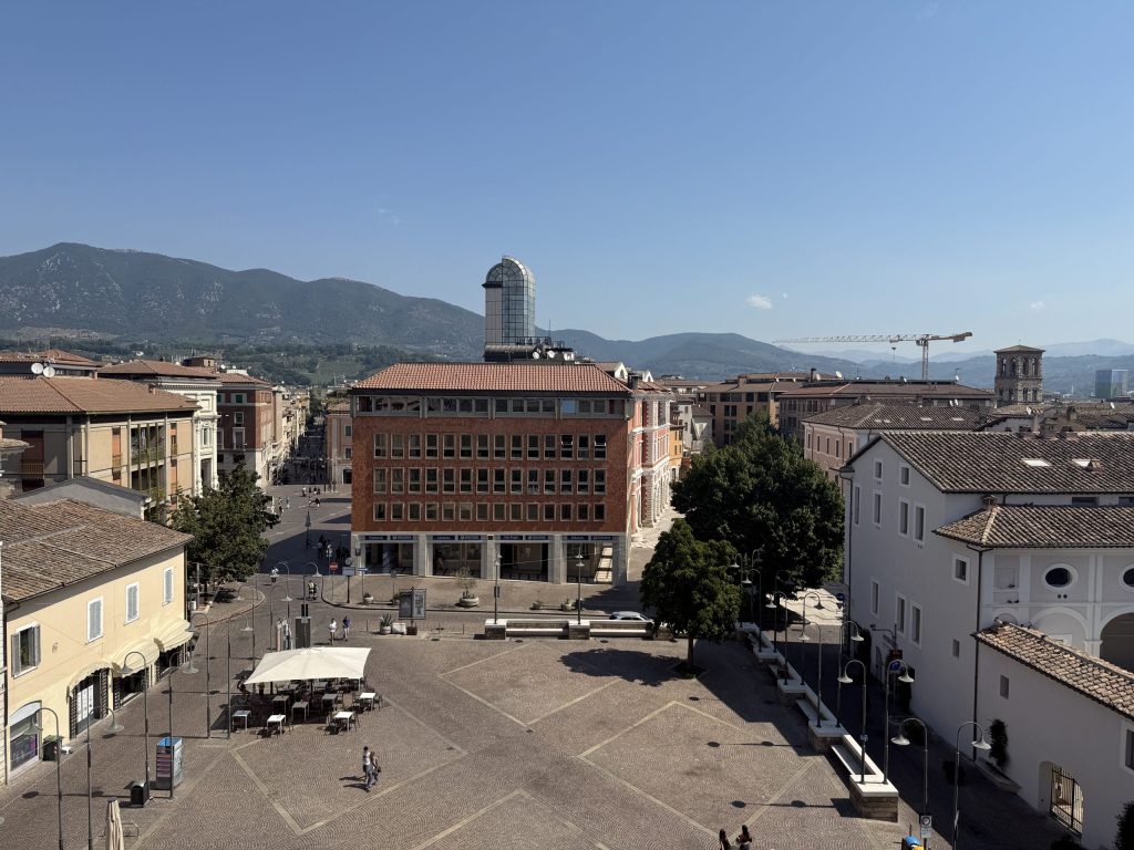 Vista panoramica di una piazza a Terni con edifici e montagne sullo sfondo, sotto un cielo sereno.