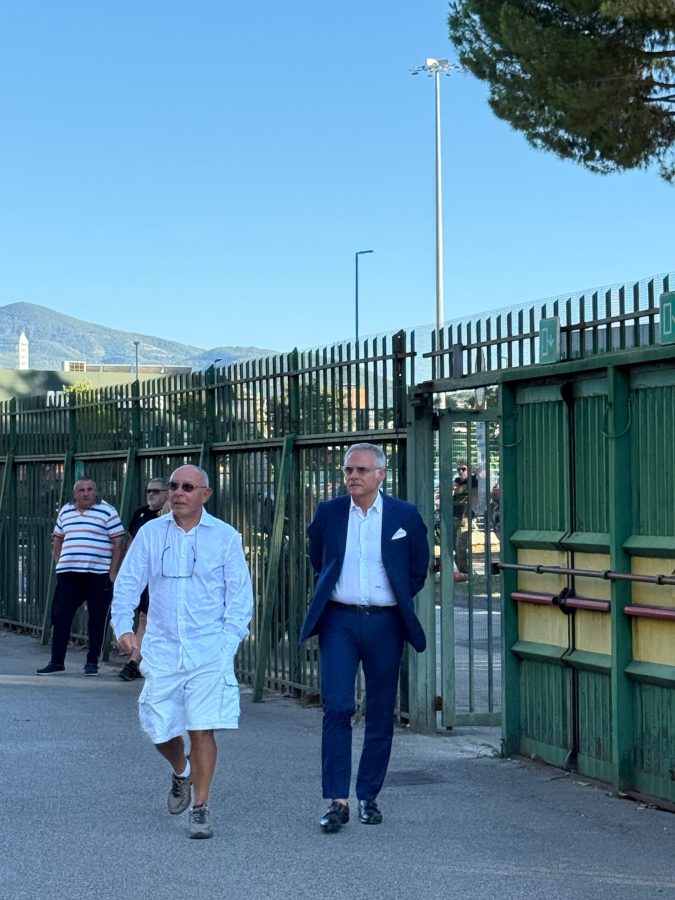 Immagine di due uomini che camminano verso l'ingresso di uno stadio, con uno in abbigliamento casual e l'altro in giacca e cravatta. Sullo sfondo si intravedono altri spettatori e una barriera verde.