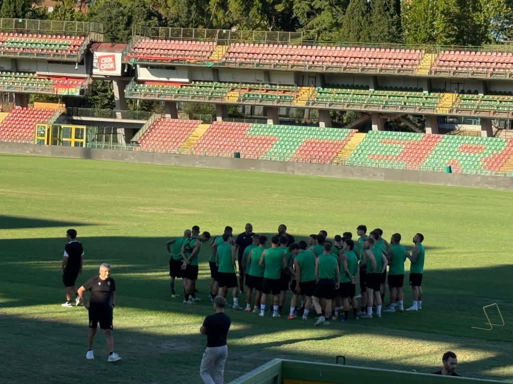 Squadra di calcio in allenamento allo stadio Libero Liberati, con atleti in maglietta verde raccolti in cerchio sotto la guida di uno staff tecnico.