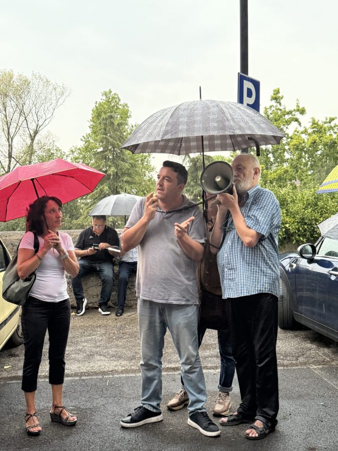 Manifestazione pacifica contro il taglio di alberi, con partecipanti sotto ombrelli che ascoltano un oratore con un megafono.