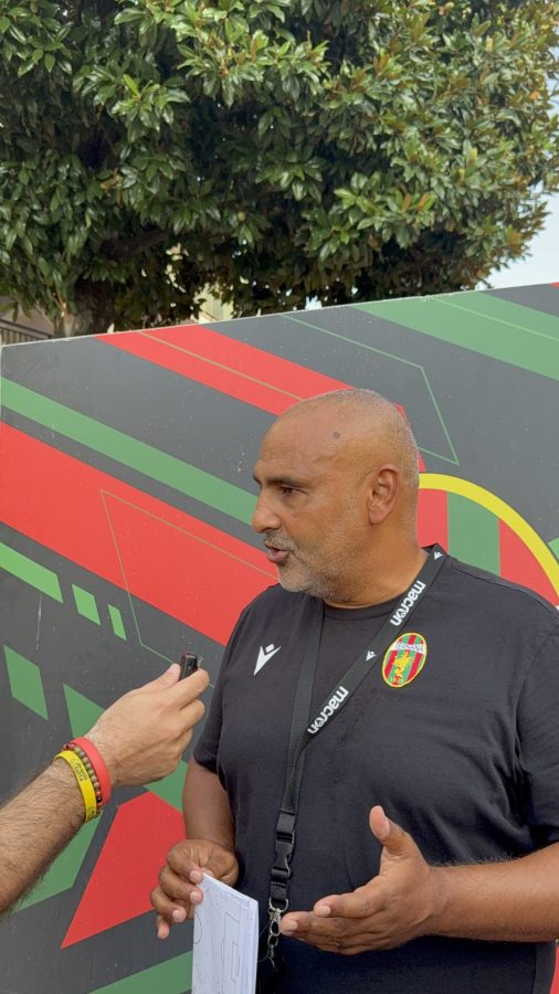 Un allenatore in maglia nera con il logo della squadra parla a un giornalista mentre tiene una cartella informativa in mano, con uno sfondo colorato di forme geometriche.