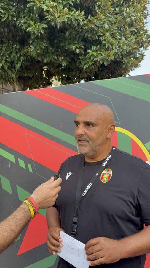 Un allenatore in maglietta nera parla a un microfono durante un'intervista, con un grande murales colorato sullo sfondo.