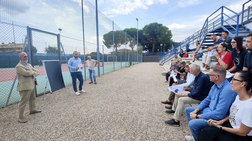 Un incontro all'aperto con un relatore che presenta un progetto, di fronte a un pubblico seduto su gradinate, in un'area sportiva.