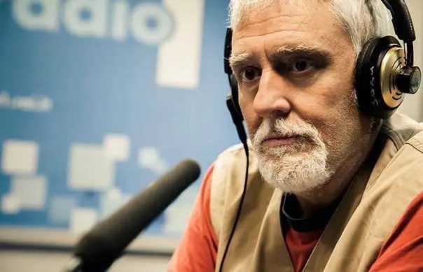 Un uomo con la barba e le cuffie sta parlando in un ambiente radiofonico. Indossa un giubbotto e sembra concentrato.