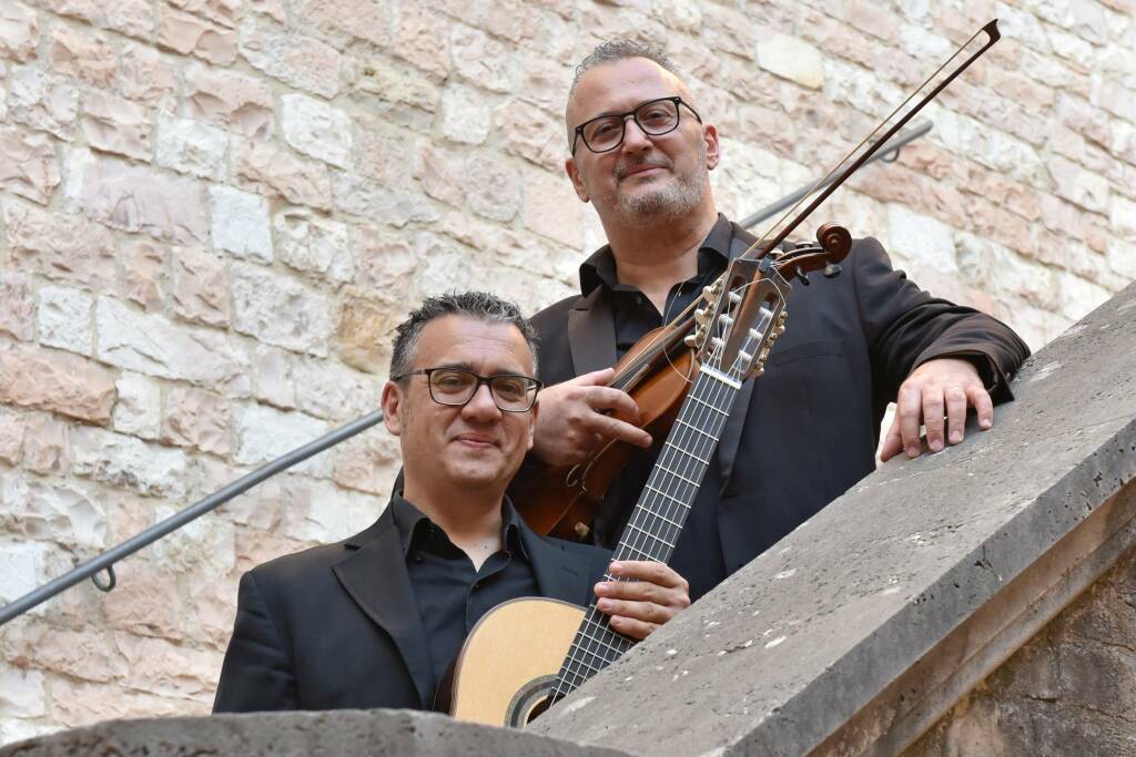Due musicisti che posano su una scala di pietra, uno con una chitarra e l'altro con un violino, davanti a un muro di pietra.