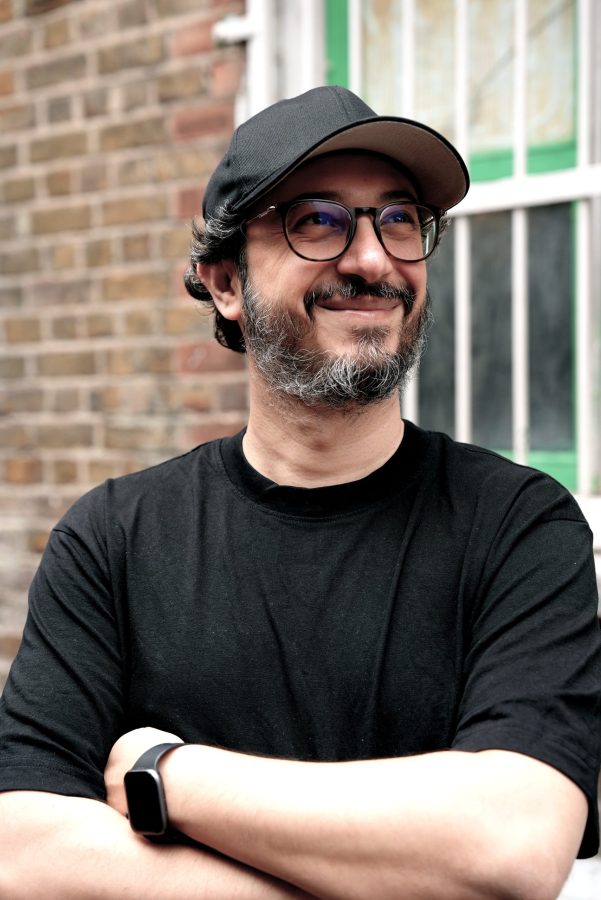 Un uomo sorridente con occhiali e cappellino nero indossa una maglietta nera, in posa con le braccia incrociate davanti a un muro di mattoni e finestre verdi sullo sfondo.