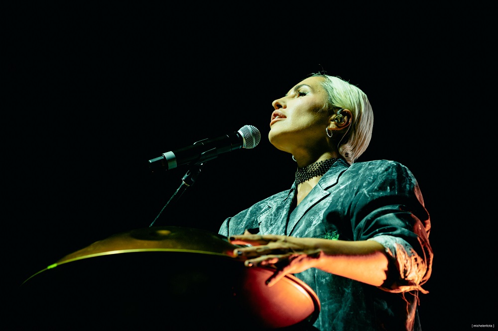 Una cantante in primo piano mentre si esibisce, con un microfono in mano e una espressione intensa, sullo sfondo scuro.