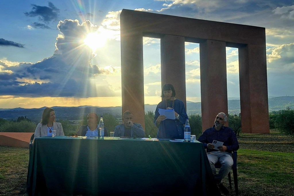 Un evento presso il Parco delle Sculture di Brufa, con persone sedute a un tavolo mentre una donna legge un documento; sullo sfondo è visibile una scultura e un paesaggio con il sole che splende attraverso le nuvole.