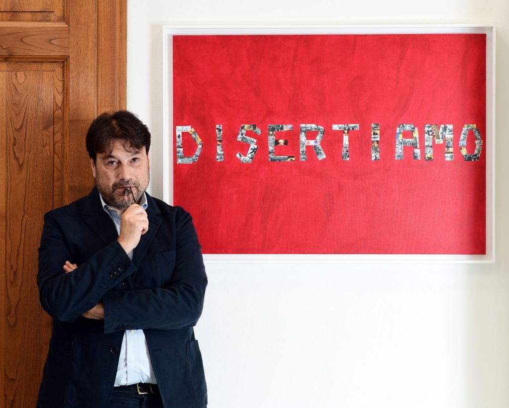 Un uomo in blazer scuro si trova di fronte a un quadro rosso con la parola 'DISERTIAMO' formata da immagini ritagliate, mentre riflette con una penna in bocca.