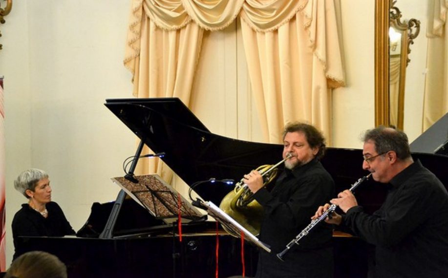 Una performance musicale dal vivo con un trio composto da un oboista, un cornovista e una pianista, sullo sfondo di un elegante ambiente interno con tende e specchi.