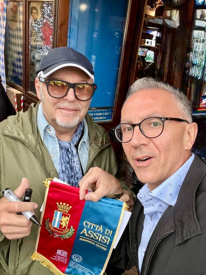 Due uomini si scattano un selfie mentre tengono in mano il gonfalone della città di Assisi. Sullo sfondo si vede un locale decorato.