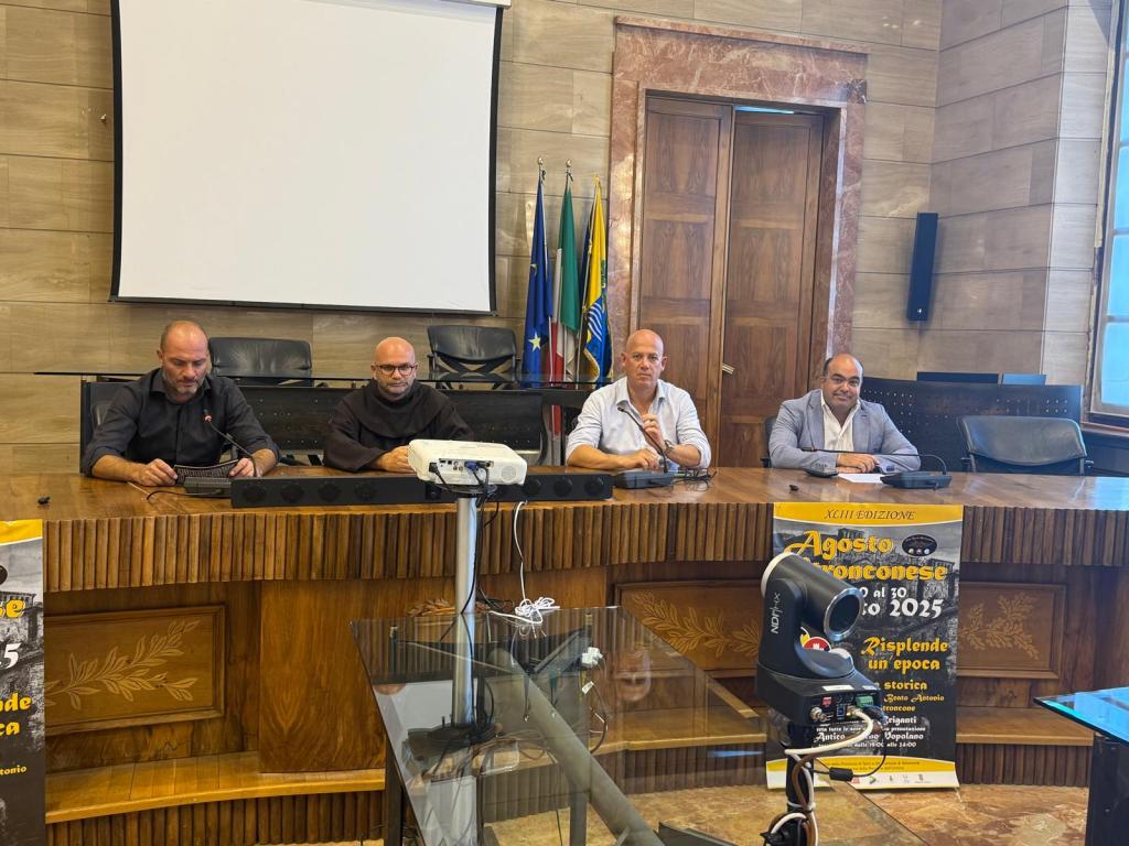 Conferenza stampa per la presentazione della 43esima edizione dell'Agosto Stronconese, con relatori seduti a un tavolo davanti a un grande schermo e striscioni informativi.