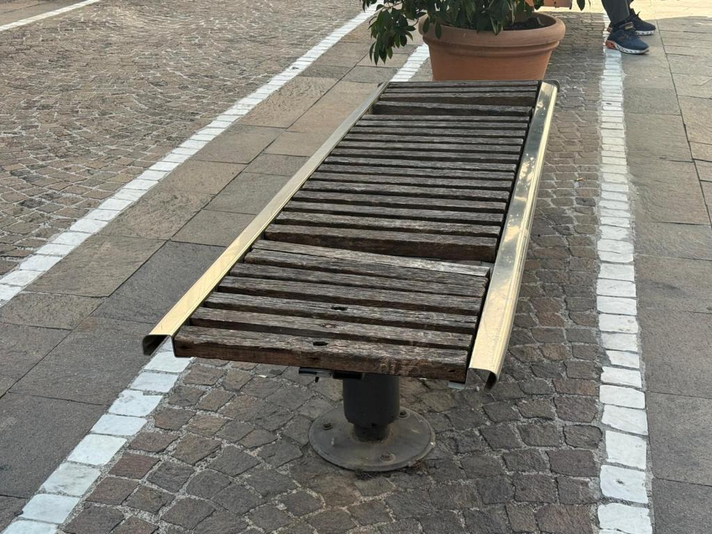 A wooden bench with metal edges, placed on a cobblestone pavement, next to a potted plant.