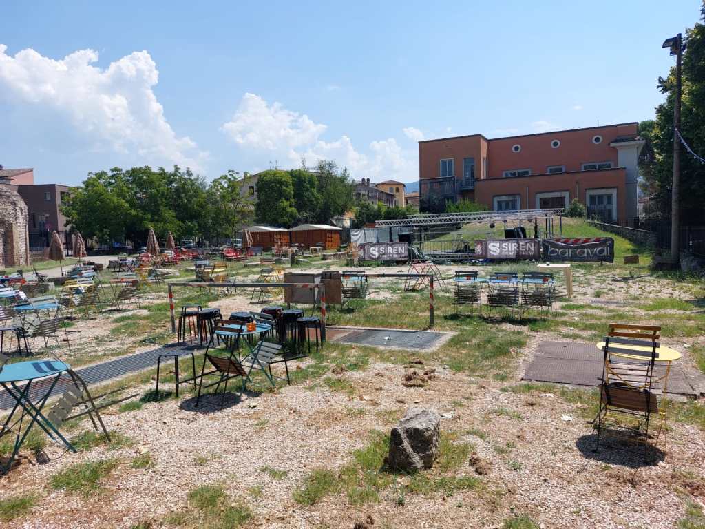 A view of a vacant outdoor space with scattered chairs and tables, surrounded by greenery and buildings under a blue sky.