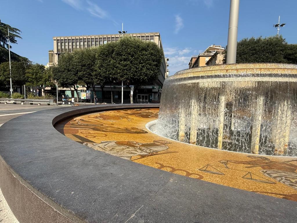 A wide view of a decorative fountain with water cascading over it, surrounded by a patterned stone base featuring artistic designs, set in an urban area with trees and buildings in the background under a clear blue sky.