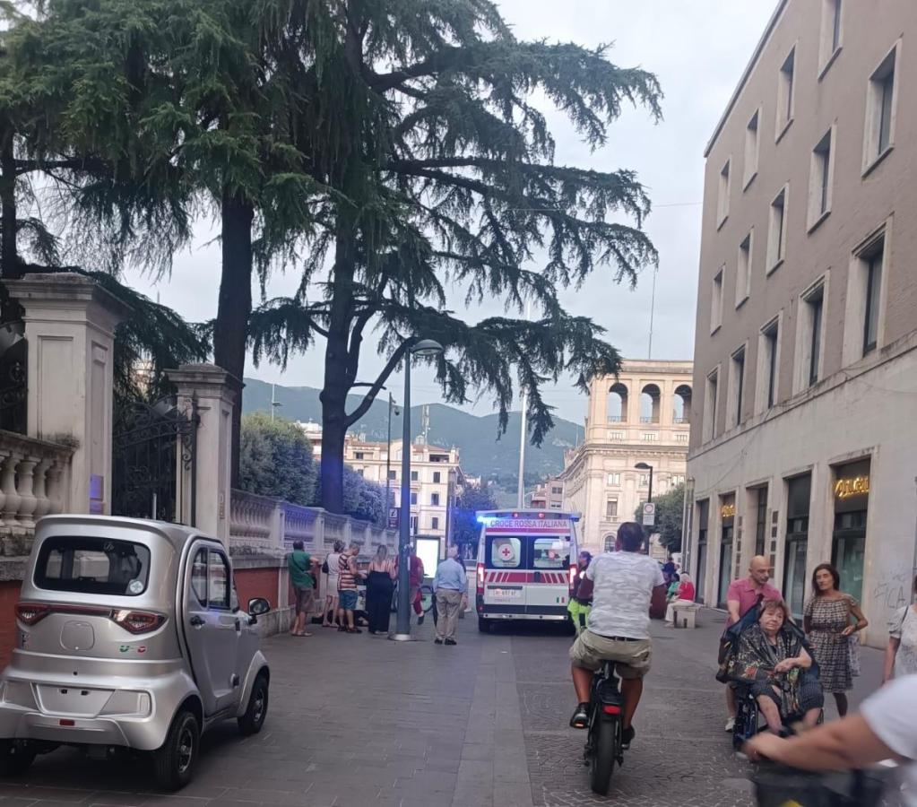A street scene in a city with a silver small car parked on the left, an ambulance with its lights on in the distance, and a group of people gathered nearby. Some individuals are walking, while one person rides a bicycle. There are buildings and trees in the background.