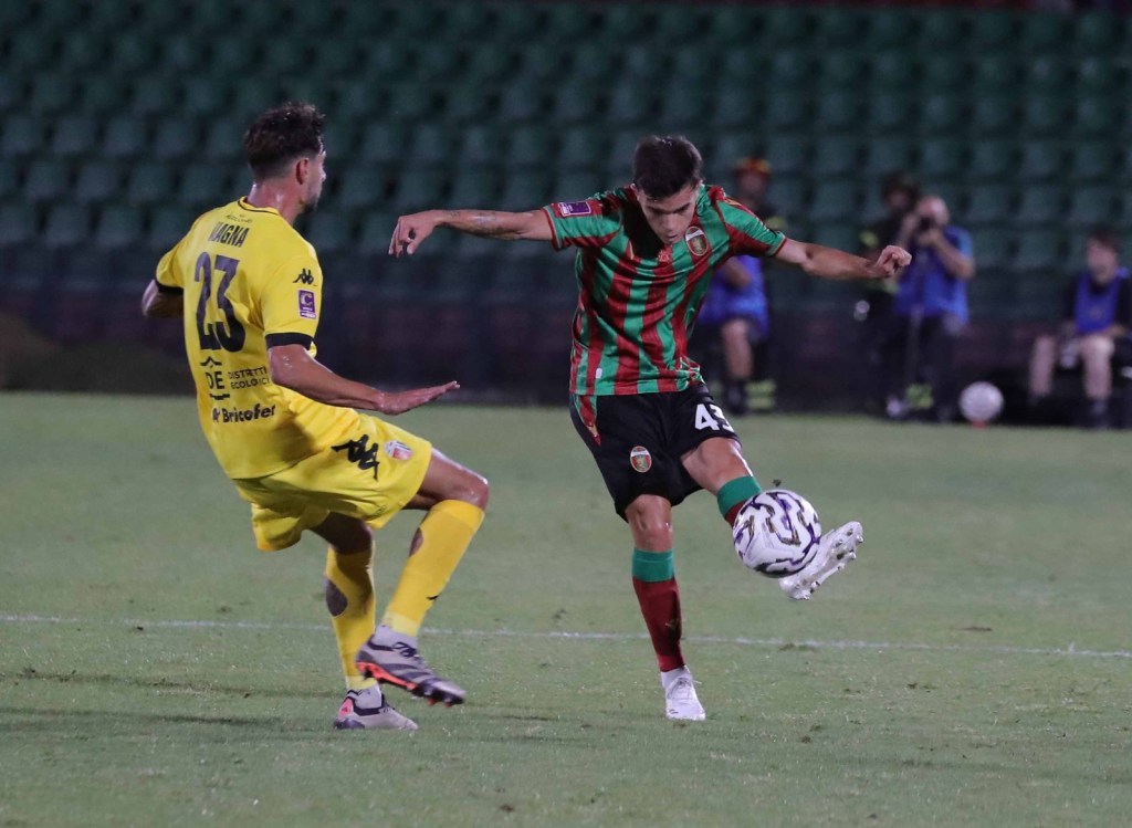 A soccer player in a red and green striped jersey shoots the ball while being challenged by an opponent in a yellow jersey during a match.