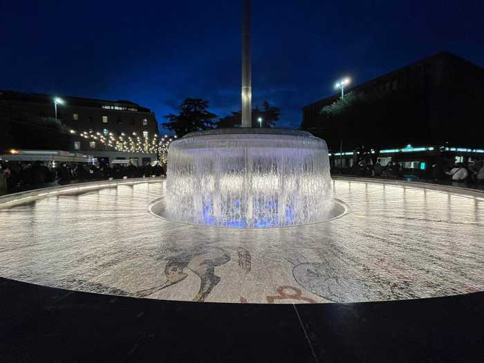 Fontana monumentale dello Zodiaco illuminata di blu durante la notte, con persone in lontananza e luci festive sullo sfondo.