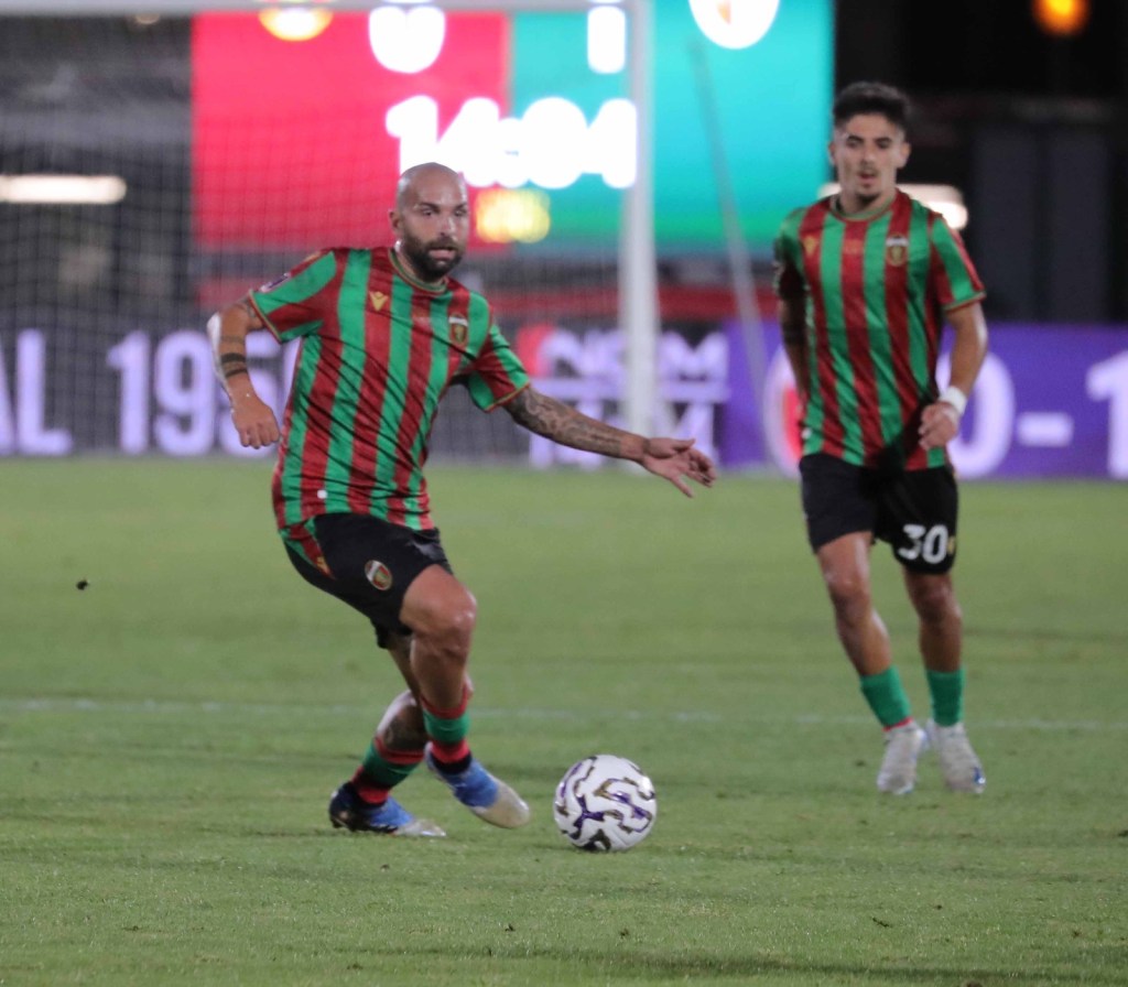 Two soccer players on the field in action during a match, with one player about to kick the ball. Both players are wearing red and green striped jerseys.