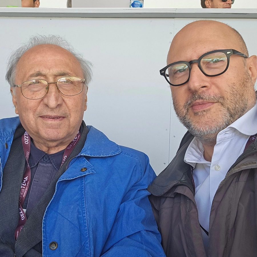 Due uomini sorridenti in una tribuna, uno con occhiali e giacca azzurra, l'altro con occhiali e giacca scura.