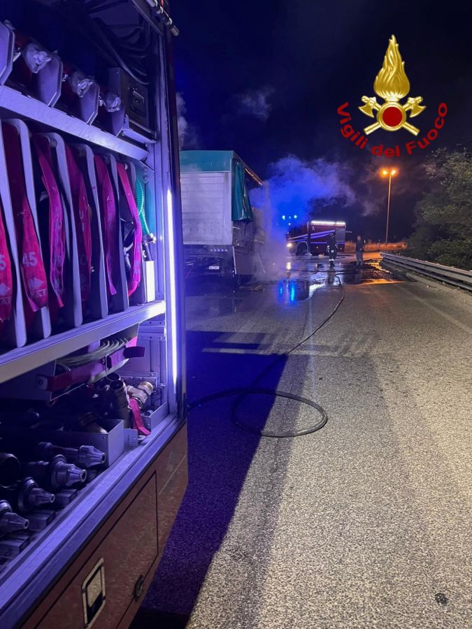 Un camion con rimorchio in fiamme lungo la SS77 Var, con vigili del fuoco che stanno spegnendo l'incendio. Notte, strada bagnata e illuminazione dei veicoli di emergenza.