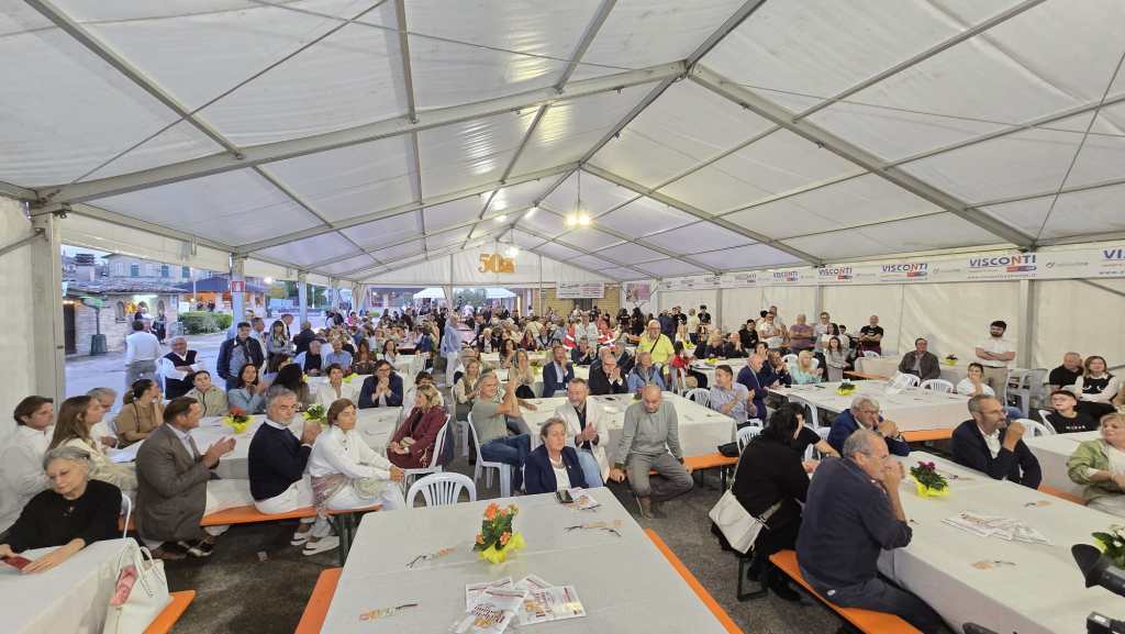 Un'immagine di una grande tenda durante l'inaugurazione della Sagra della porchetta di Costano, con un folto pubblico seduto ai tavoli, mentre alcuni partecipanti applaudono. La tenda è decorata con fiori e striscioni.