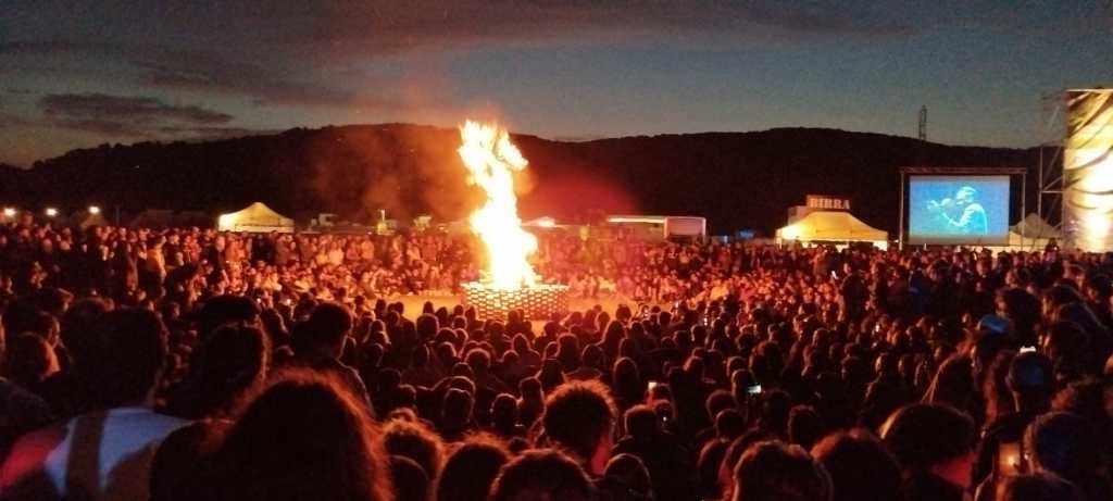 Una folla che assiste a un grande falò durante il Montelago Celtic Festival, sullo sfondo si vedono tende e un palco con immagini proiettate.