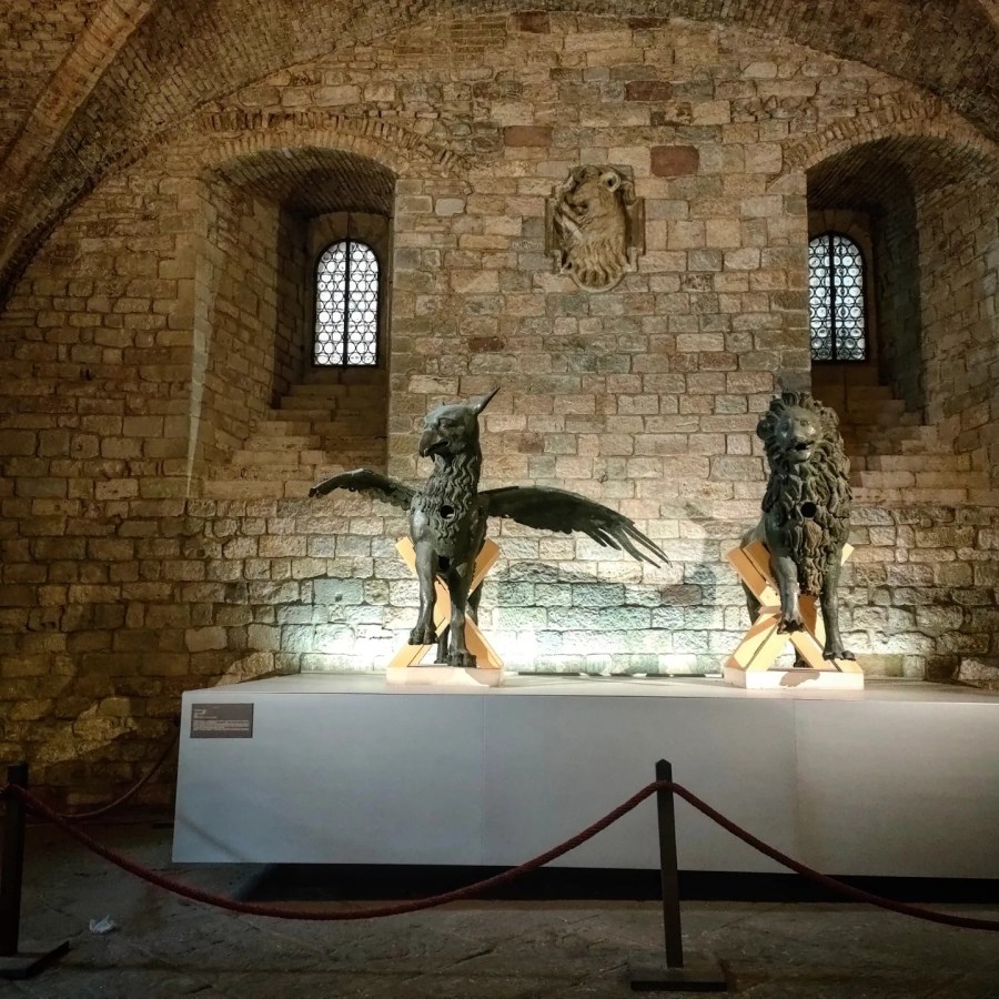 Due sculture di bronzo, un grifone e un leone, esposte in una galleria con pareti in pietra, illuminate da una luce soffusa.