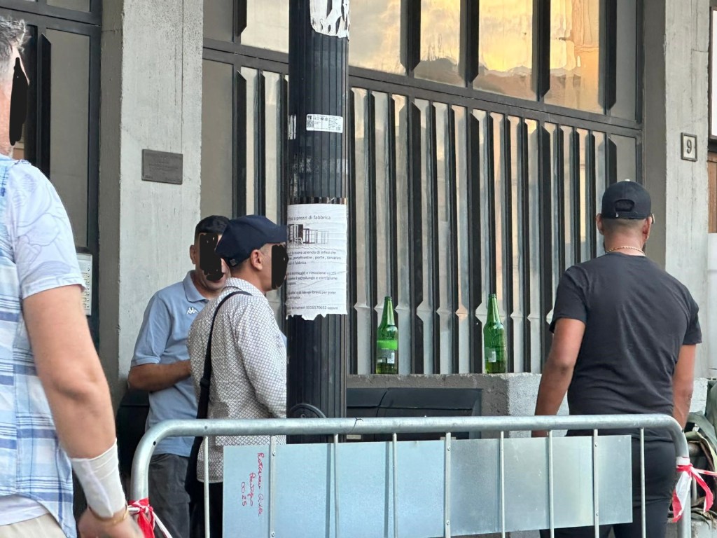 Due uomini davanti a un'attività commerciale con bottiglie di birra verdi appoggiate al muro, mentre una terza persona è in piedi, osservando.