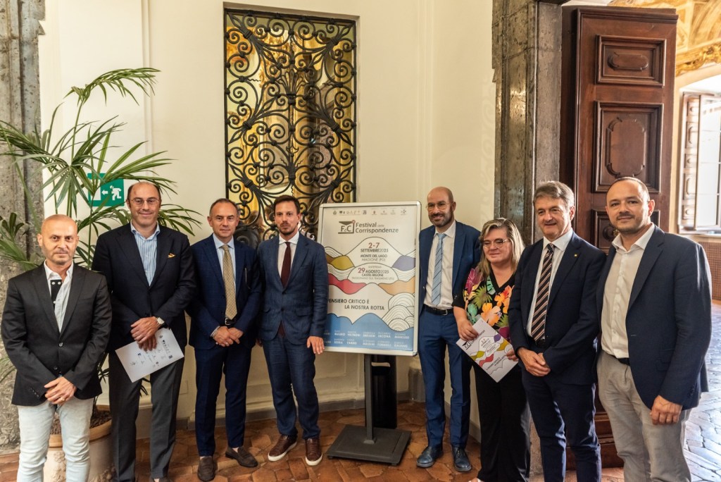 Un gruppo di otto persone riunite in una sala elegante, posizionati accanto a un poster del Festival delle Corrispondenze, con decorazioni floreali e dettagli architettonici sullo sfondo.