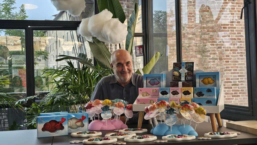 Un uomo sorridente seduto davanti a una varietà di prodotti di cioccolato, tra cui dolci a forma di nuvole e leccalecca colorati, con una decorazione di nuvole sospese sullo sfondo.