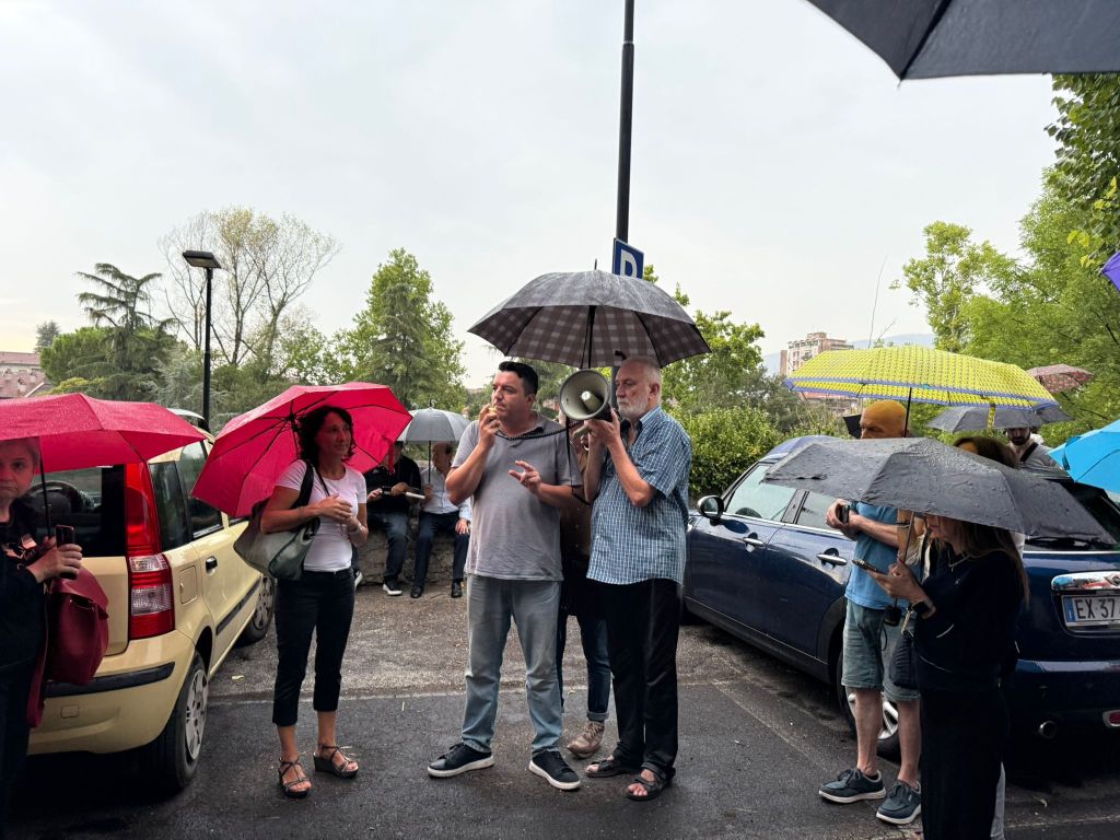 A diverse group of people standing outside under umbrellas during rainy weather, with one person using a megaphone to speak.