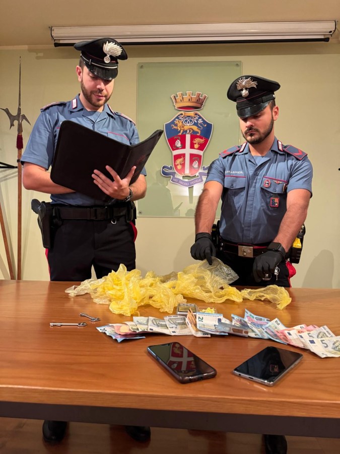 Due carabinieri esaminano materiali e denaro sequestrati su un tavolo, con un distintivo sullo sfondo.
