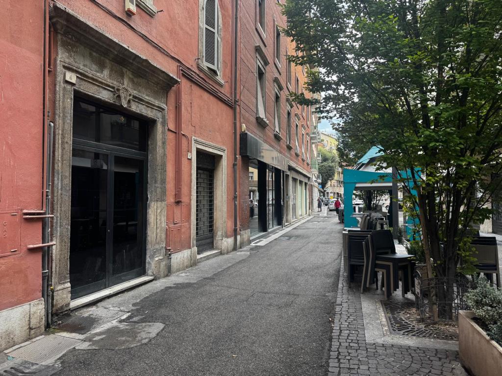 Una vista della via Goldoni a Terni, caratterizzata da saracinesche abbassate, vetrine vuote e cartelli 'affittasi'. L'atmosfera è desolante, con pochi passanti e tavoli all'aperto non utilizzati.