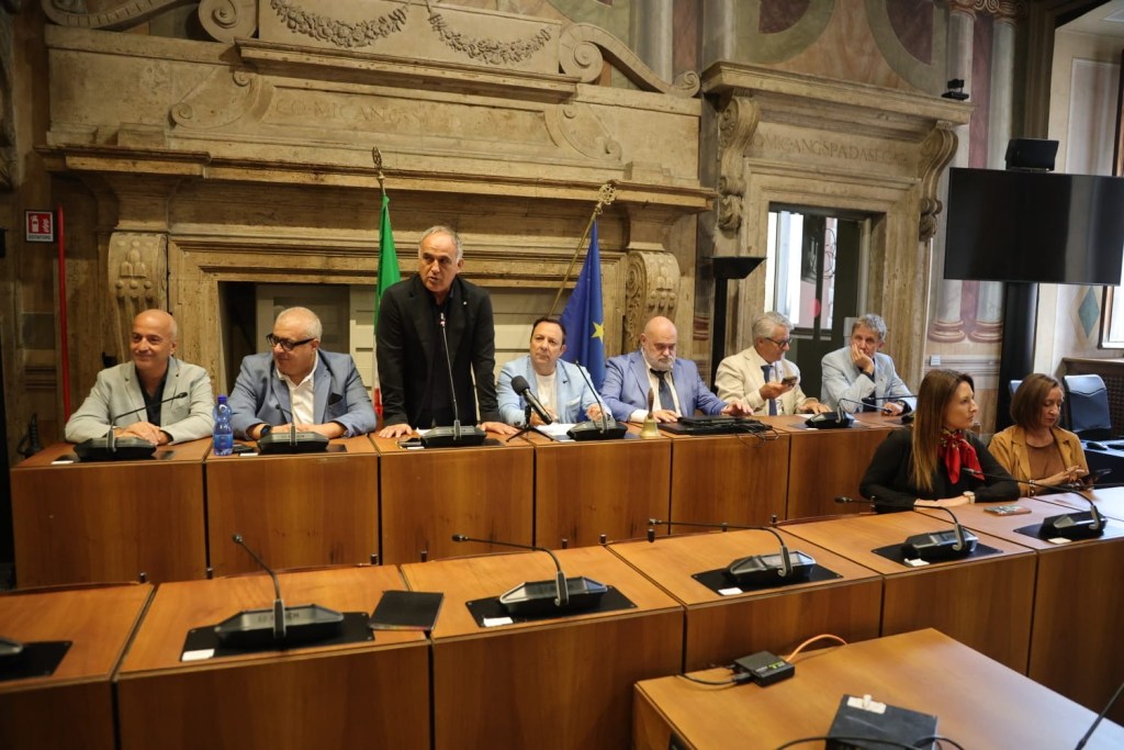 A group of eight people seated at a long table in a formal meeting room, with an Italian flag and an EU flag in the background.
