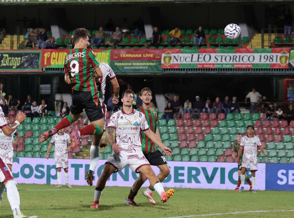 Un giocatore in maglia verde e rossa salta per colpire il pallone durante una partita di calcio, mentre altri giocatori in maglie bianche lo osservano, sullo sfondo uno stadio affollato.