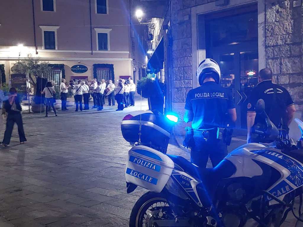 A nighttime street scene featuring a group of musicians playing instruments in a square, with several police officers observing. The police are on motorcycles, and the setting includes buildings with illuminated windows.