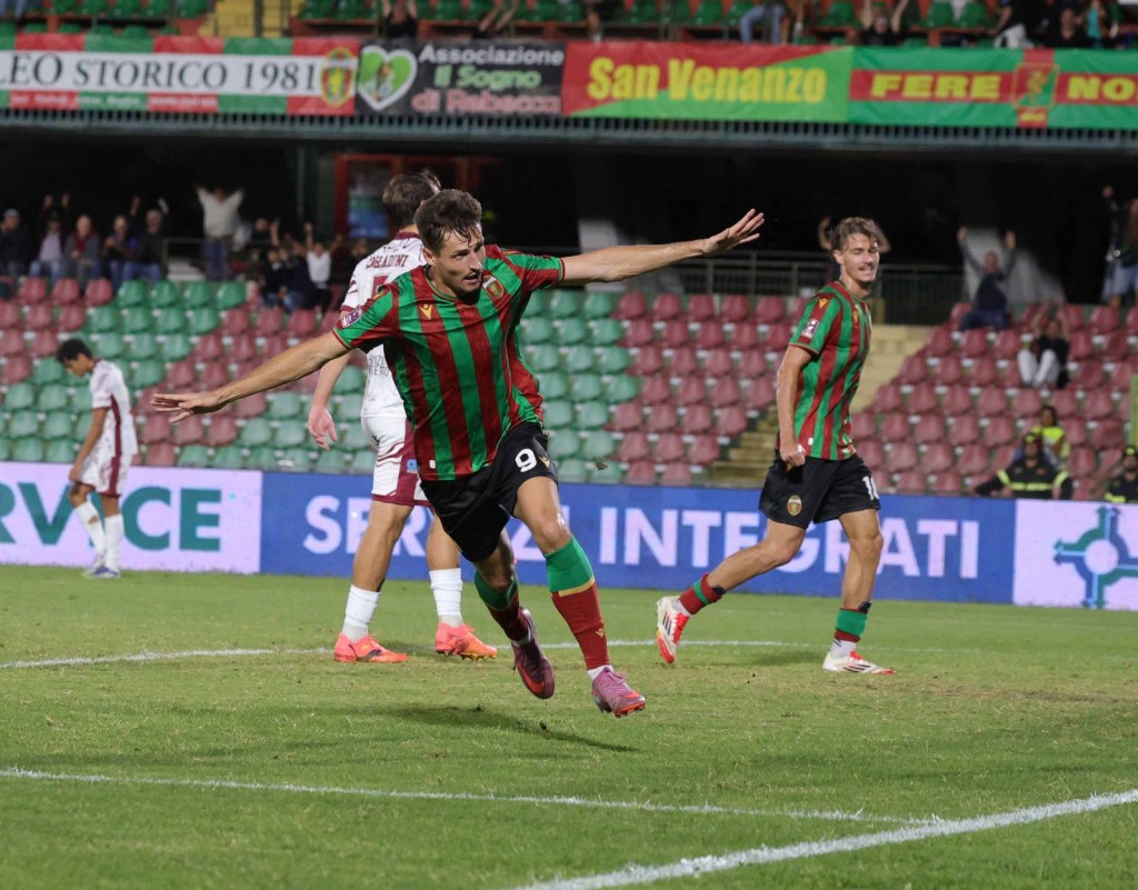 Giocatore che celebra un gol durante una partita di calcio, indossando una maglia a strisce verdi e rosse. Sullo sfondo, alcuni spettatori e un altro giocatore avversario.