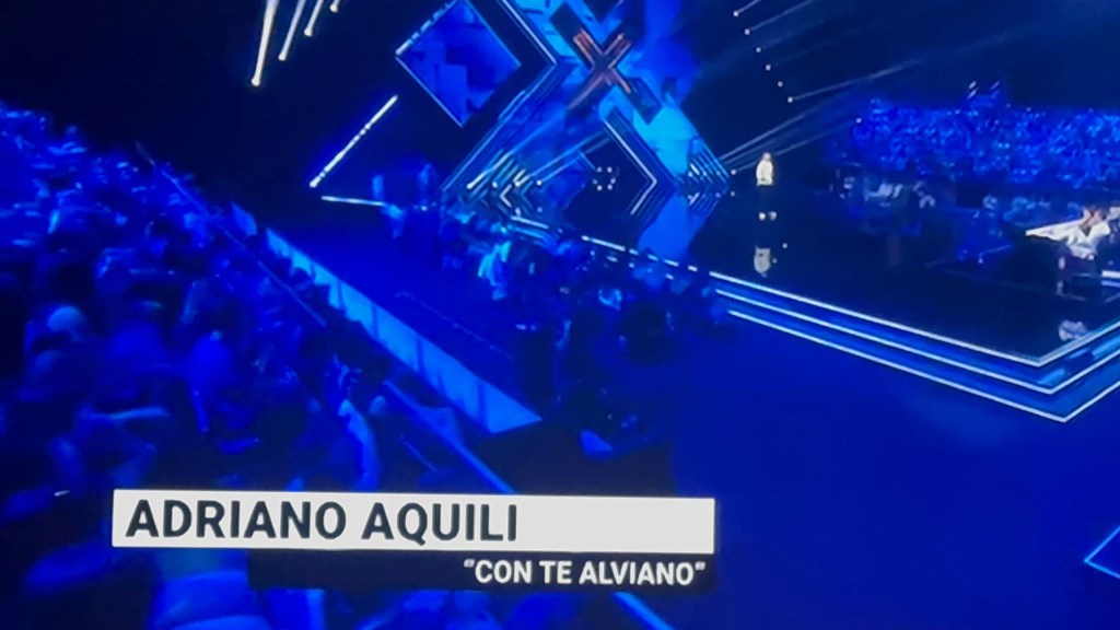 Stage view featuring performer Adriano Aquili, with blue lighting and an audience in the background.