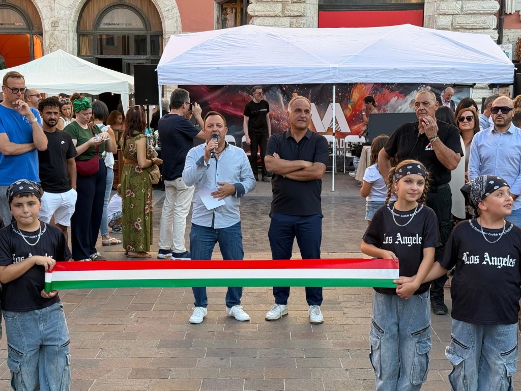 A group of people at an outdoor event, with two children in front holding a tricolor ribbon. A man with a microphone is speaking to the crowd, while others watch attentively.