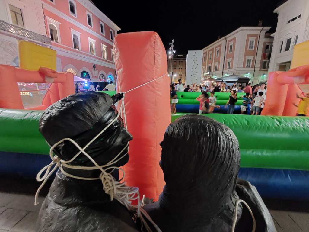 View of a lively nighttime festival with inflatable attractions, featuring two black statues in the foreground and a crowd enjoying the event in the background.