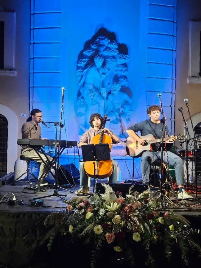 Three musicians performing on stage, with a pianist on the left, a cellist in the middle, and a guitarist on the right, in front of a lit backdrop featuring a stone sculpture.