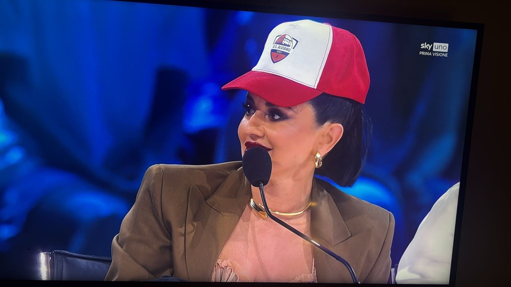 A woman wearing a red and white cap and a brown blazer sits at a table, looking to her left with a microphone in front of her.