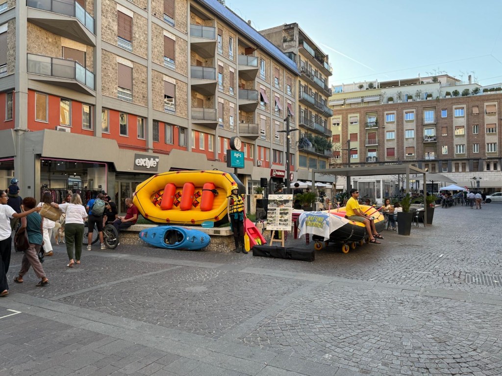 A lively street scene featuring people walking past shops, with colorful kayaks and inflatable boats displayed prominently in the square.