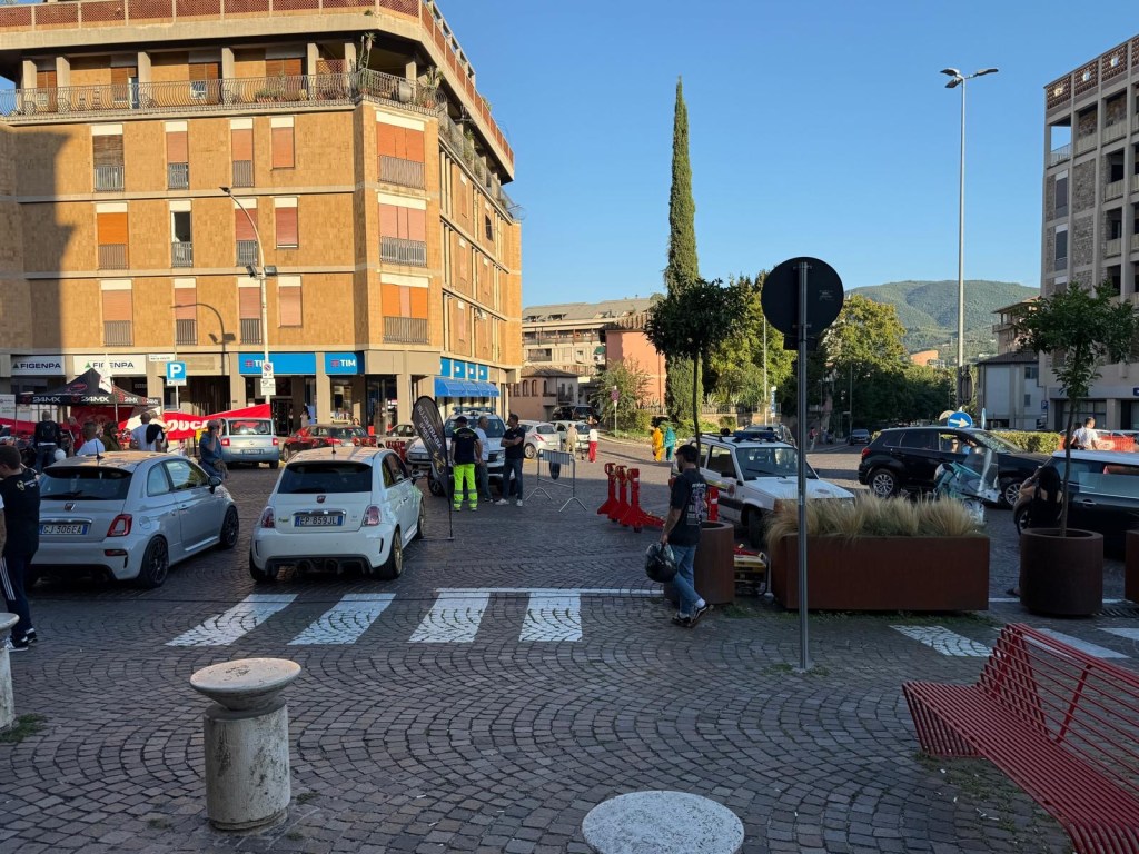 A busy street scene depicting several cars parked along a cobblestone road, with people walking and interacting. In the background, a tall building and trees are visible against a clear blue sky.